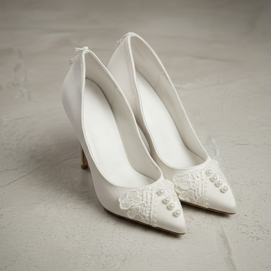 Pair of white wedding shoes with lace and pearl details on a light gray background